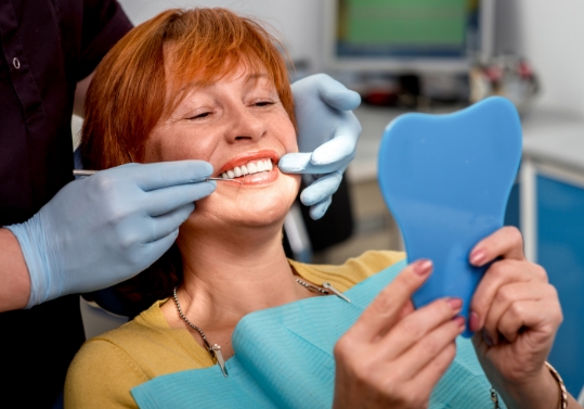 Older redheaded woman looking at her smile in mirror while sitting in dental chair